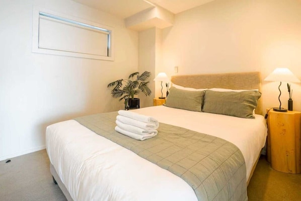 A neatly made bed with a green bedspread and white linens, two bedside lamps, a potted plant, and a wall-mounted light fixture.