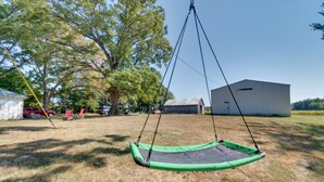 Ein Trampolin auf einer grünen Wiese mit einem Bauernhof und Bäumen im Hintergrund.
