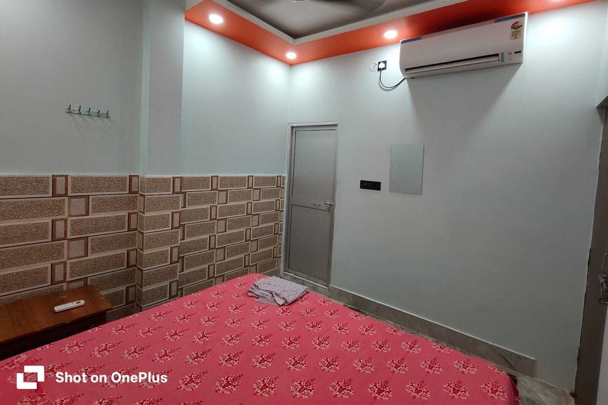 Un dormitorio con cabecera de ladrillo, una cama con colcha floral roja, un equipo de aire acondicionado y un ventilador de techo.
