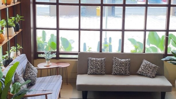 A cozy room with a sofa, a chair, a small table, and a hanging lamp. There are potted plants on shelves and a large window with a view of the outside. - Hotel Setia Budi Madiun (Madiun)