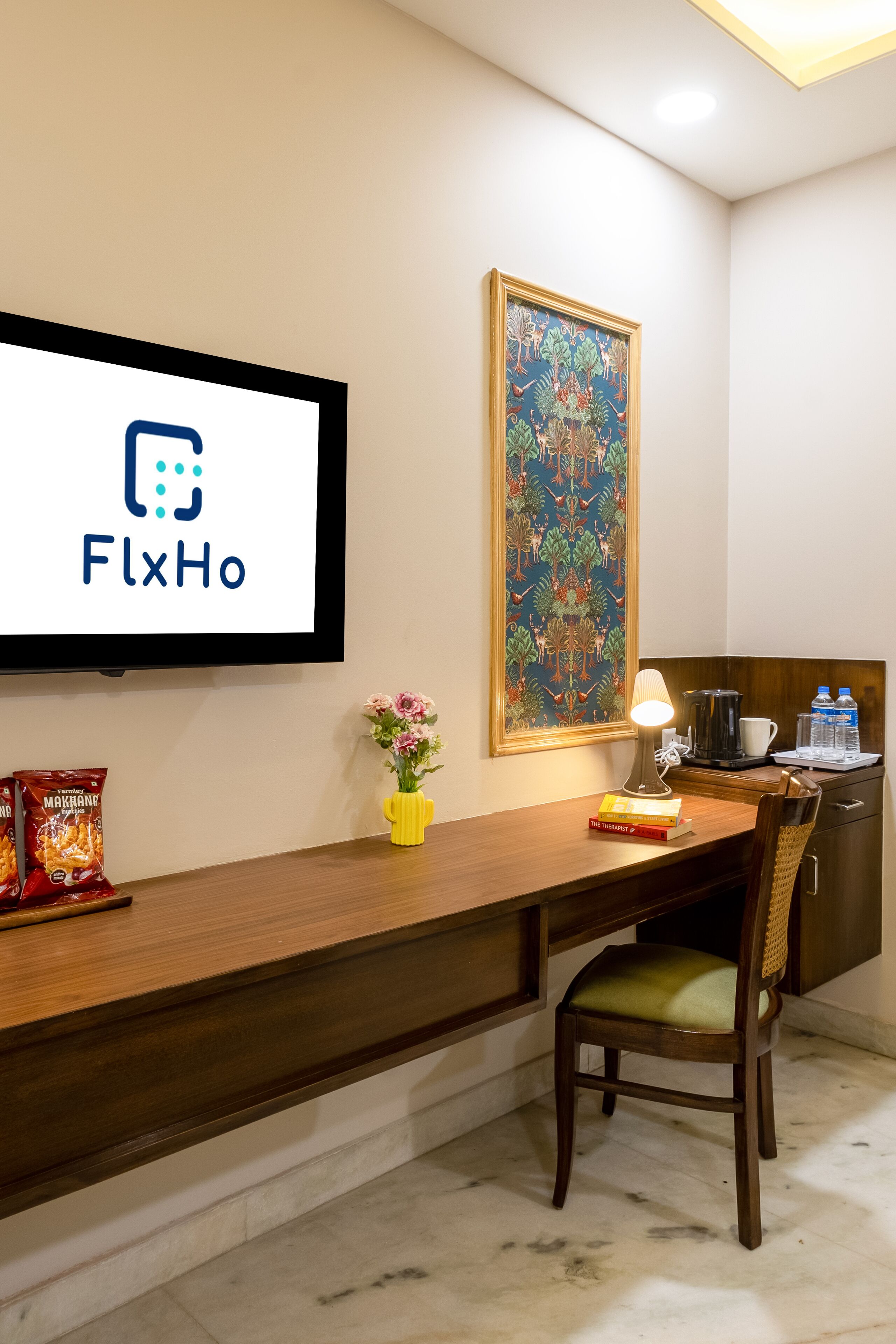 Een hotel receptie met een bureau, stoel, een ingelijste kunstwerk, een televisiescherm waarop het logo 'FlxHo' staat en een koffieapparaat.