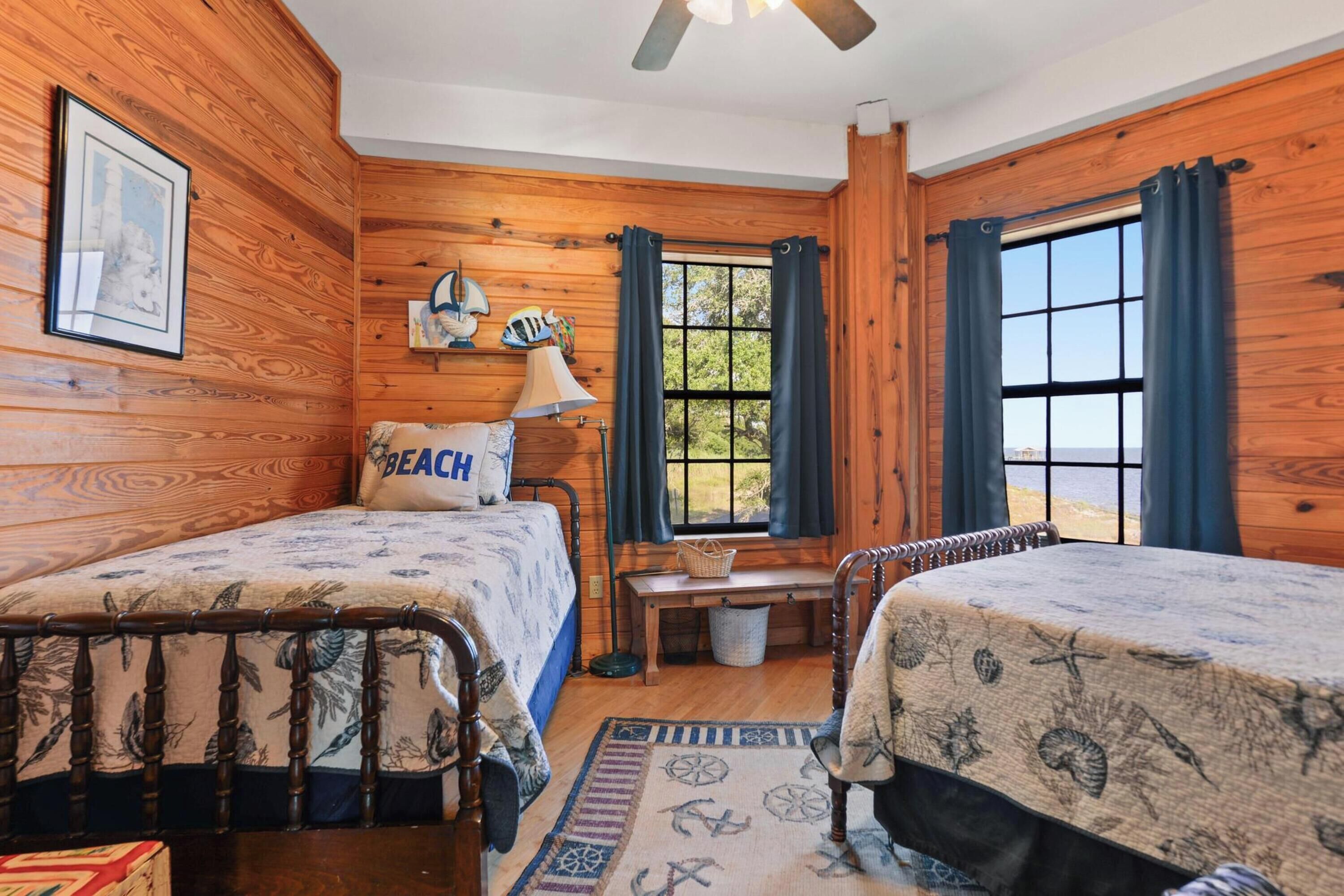 A cabin-style room with two beds, a wooden ceiling fan, a window with curtains, and a view of the outdoors.