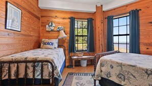 A cabin-style room with two beds, a wooden ceiling fan, a window with curtains, and a view of the outdoors.
