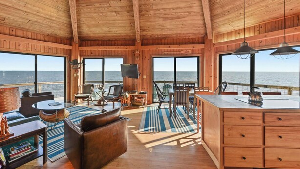 Een houten chalet met een groot balkon dat uitkijkt over de oceaan, gemeubileerd met een bank, stoelen, een eettafel en een keukenhoek.