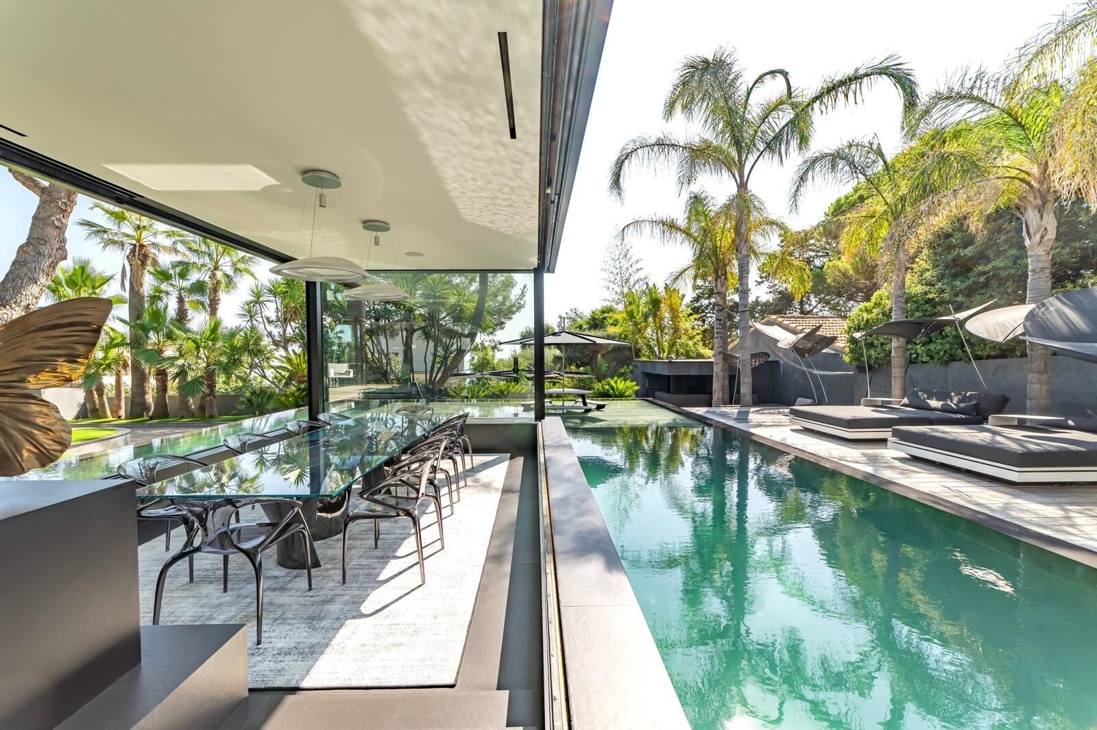 A modern outdoor pool area with a glass table and chairs, surrounded by palm trees and a view of the pool.