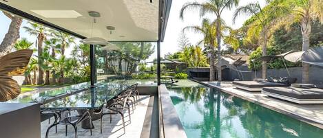 A modern outdoor pool area with a glass table and chairs, surrounded by palm trees and a view of the pool.