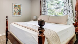 A bedroom with a large bed, wooden headboard, a window with patterned curtains, and a wall-mounted abstract painting.