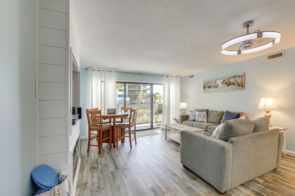 A living room with a sofa, a dining table with chairs, a ceiling light, and a view through a glass door. - Direct Beach Access! Walkable Condo on Ocean Blvd (North Myrtle Beach)