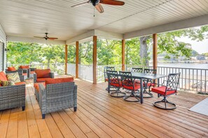 A covered outdoor deck with wicker furniture and red cushions, a dining table with chairs, and a view of a lake. - Boat, Fish & Explore! Gem on Lake of the Ozarks (Roach)