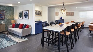 A modern dining area with a long table and multiple chairs, a sofa with colorful cushions, and a kitchen area with blue cabinets.