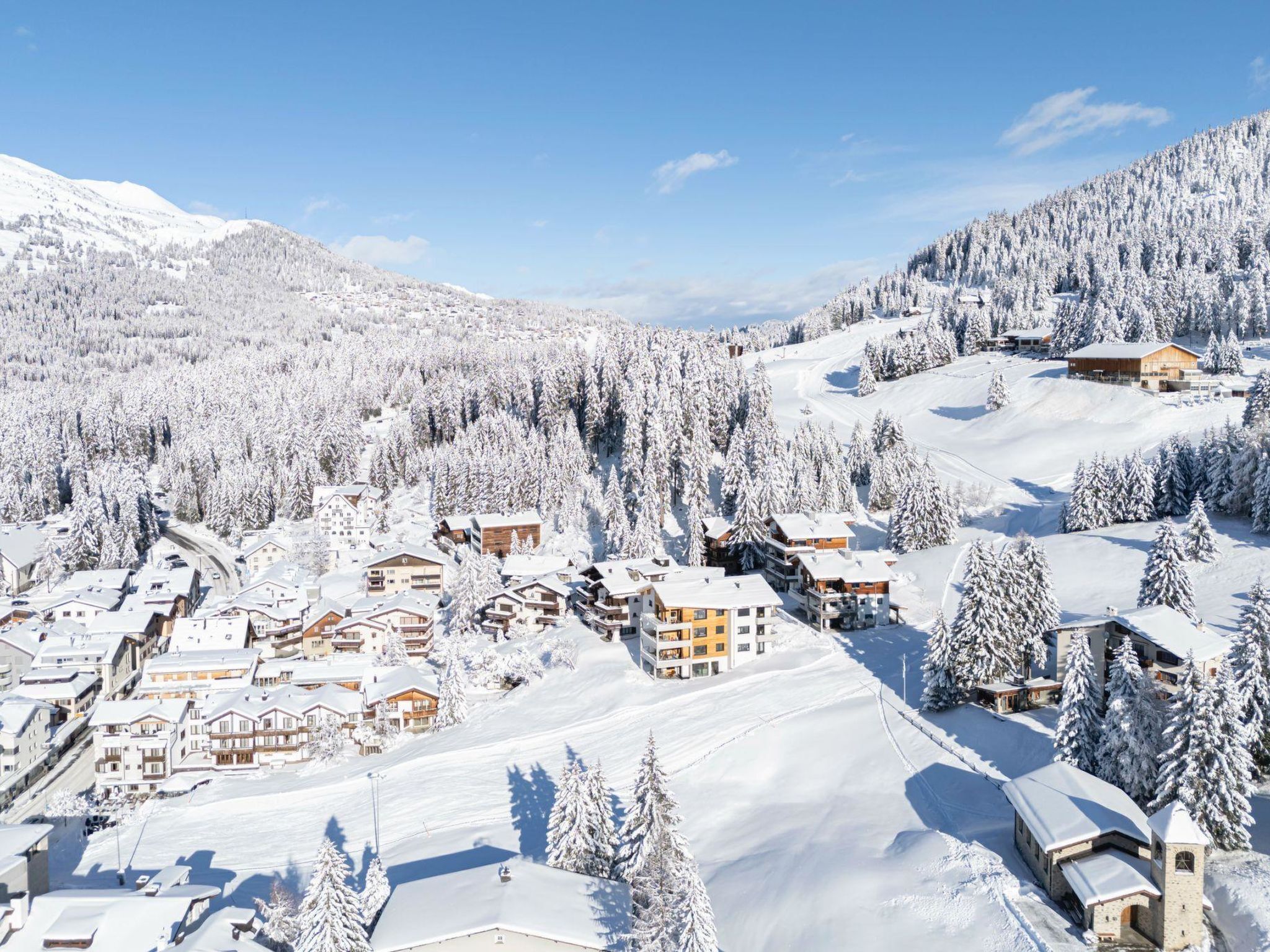 Un centro turístico en la montaña, cubierto de nieve, con varios edificios, incluyendo alojamientos y una iglesia, rodeado de bosques densos y pistas de esquí.