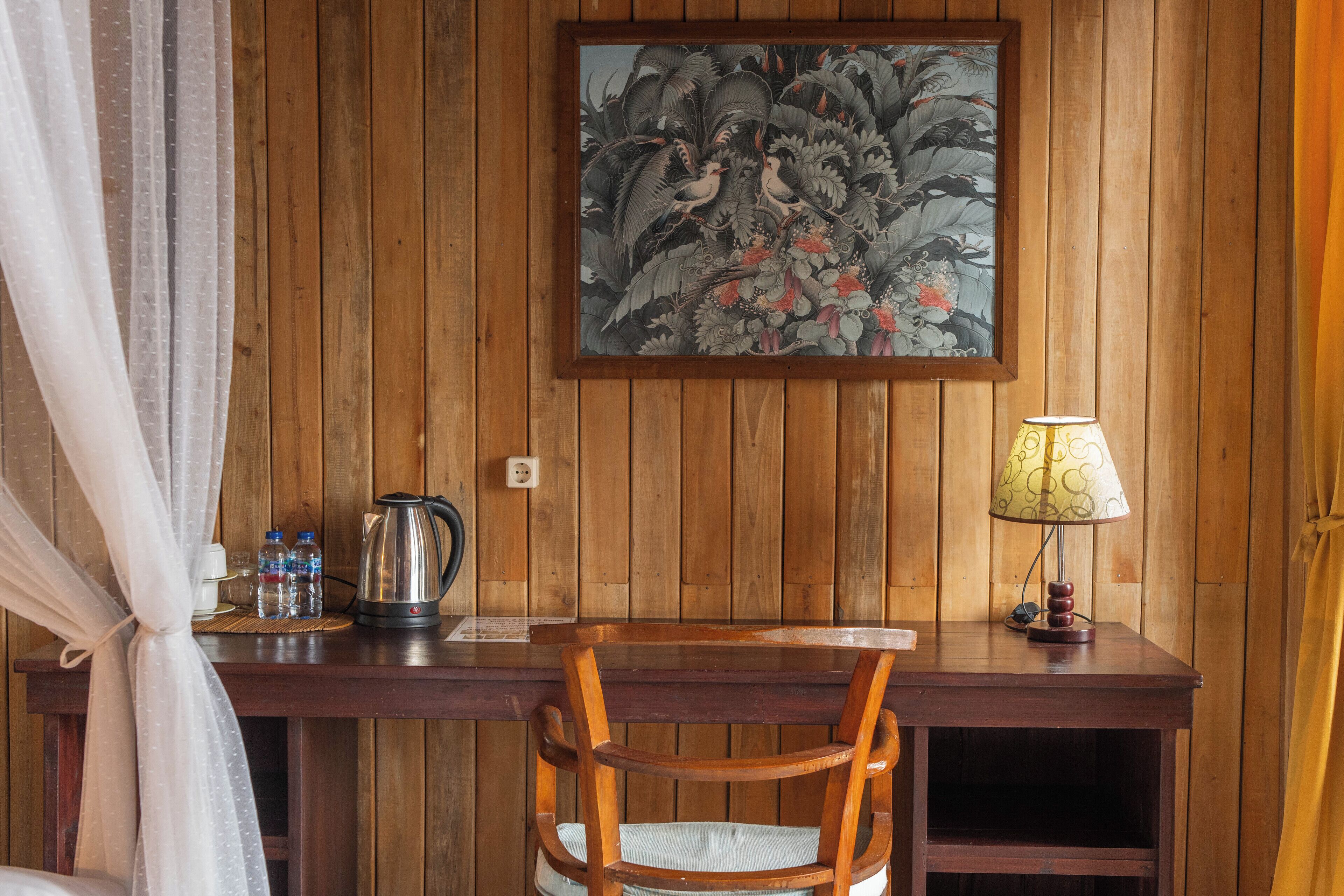 A wooden desk with a chair, a framed painting, a lamp, and a kettle.