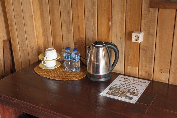 Una mesa de madera con una taza de café blanca, dos botellas de agua y una tetera eléctrica plateada. Hay un mantel individual sobre la mesa, y se ve un letrero con detalles de la propiedad.