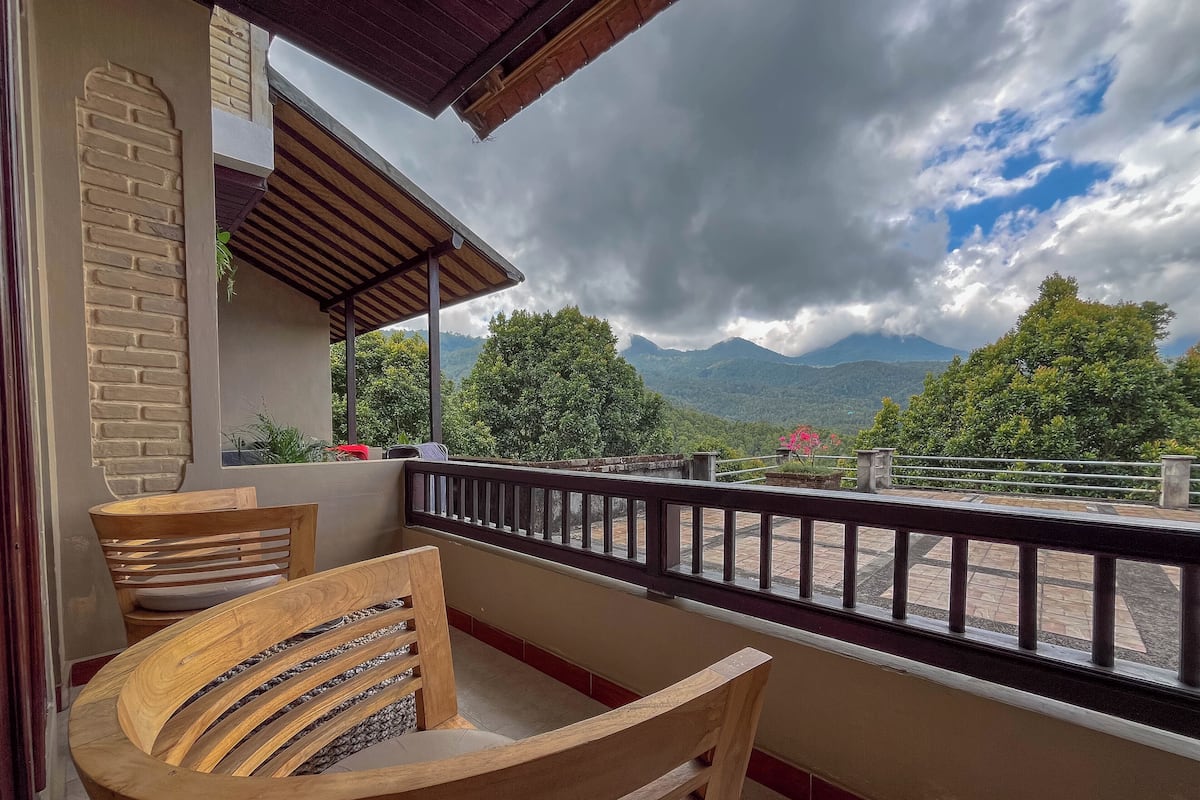 Un balcon avec des chaises en bois, une vue sur des collines verdoyantes et des montagnes, et un ciel nuageux.