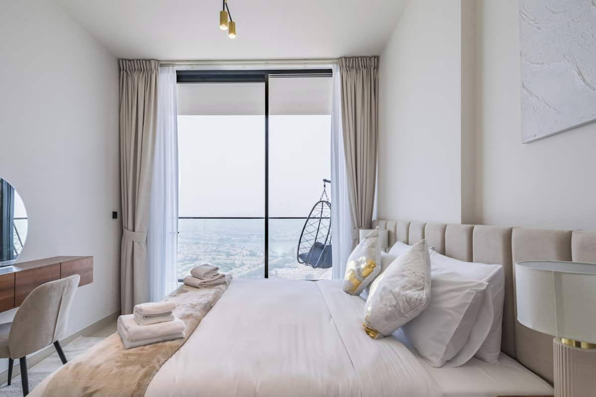 A modern hotel room with a large bed, a desk, and a view of the city through floor-to-ceiling windows.