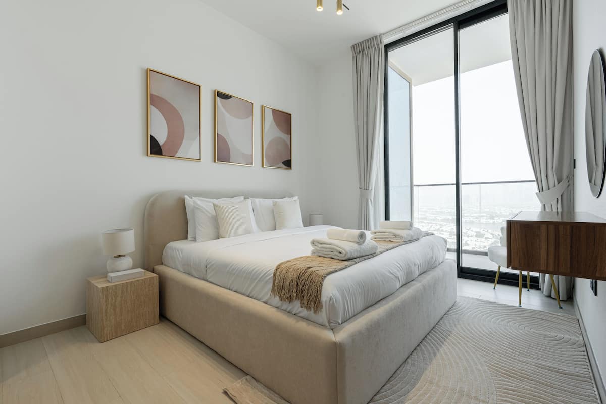 A modern bedroom with a large bed, a bedside table, a nightstand, and a view of the city through a large window.