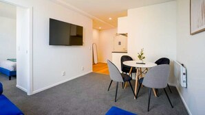 Een moderne woonkamer met een flatscreen televisie, een eethoek met een tafel en stoelen, en een zichtbare keuken op de achtergrond.