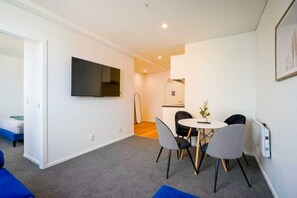 Un salon moderne doté d’un téléviseur à écran plat, d’un coin repas avec une table et des chaises, et d’une cuisine ouverte visible au fond.