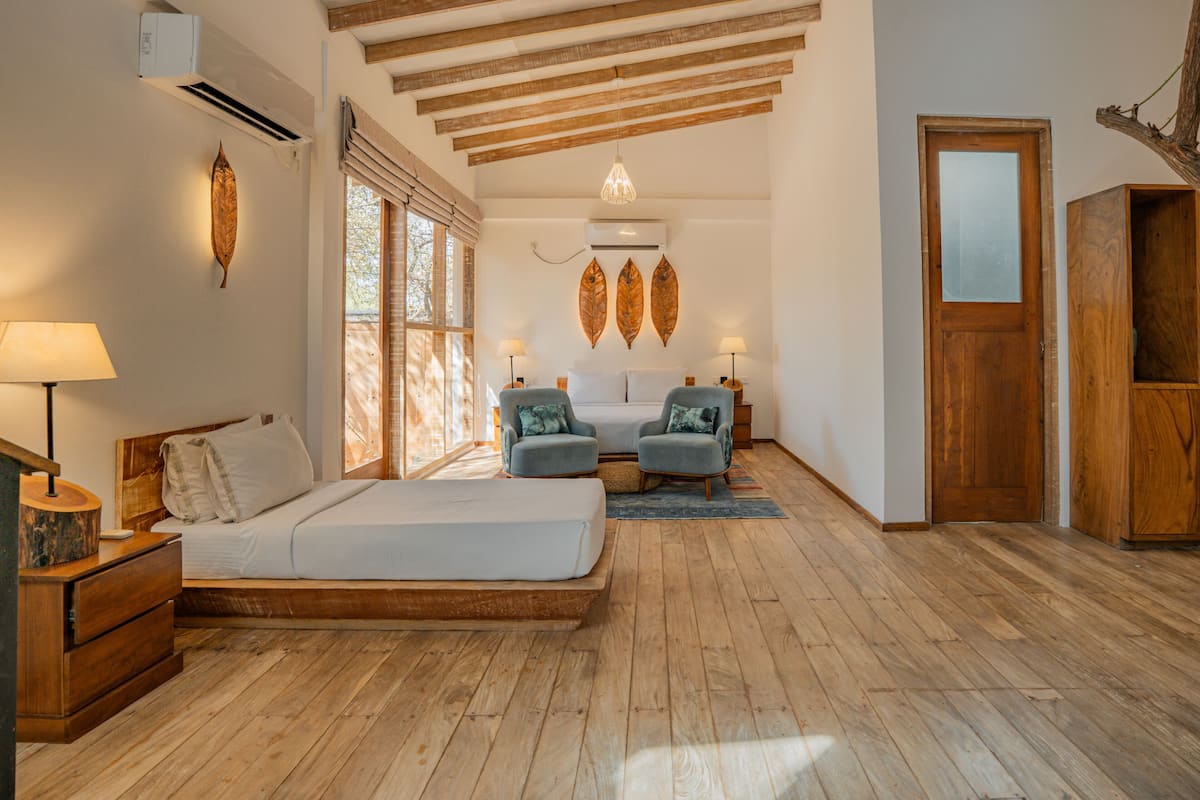 Une chambre avec un plancher en bois, un lit, deux fauteuils, une table de chevet, une lampe et une porte.
