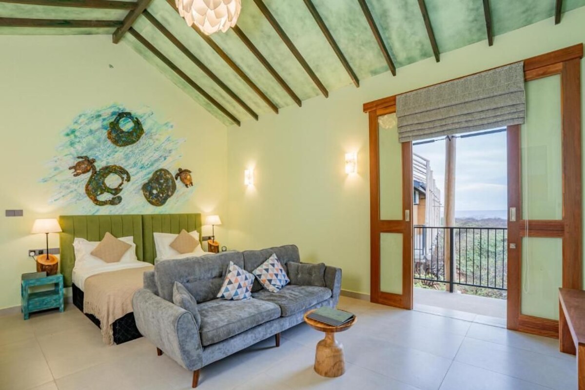 A bedroom with a bed, a gray sofa, a small table, a wall with a decorative piece, and a balcony with a view.