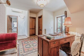 Una habitación con un escritorio de madera, libros, una silla roja, un tapete estampado y una ventana con vistas.