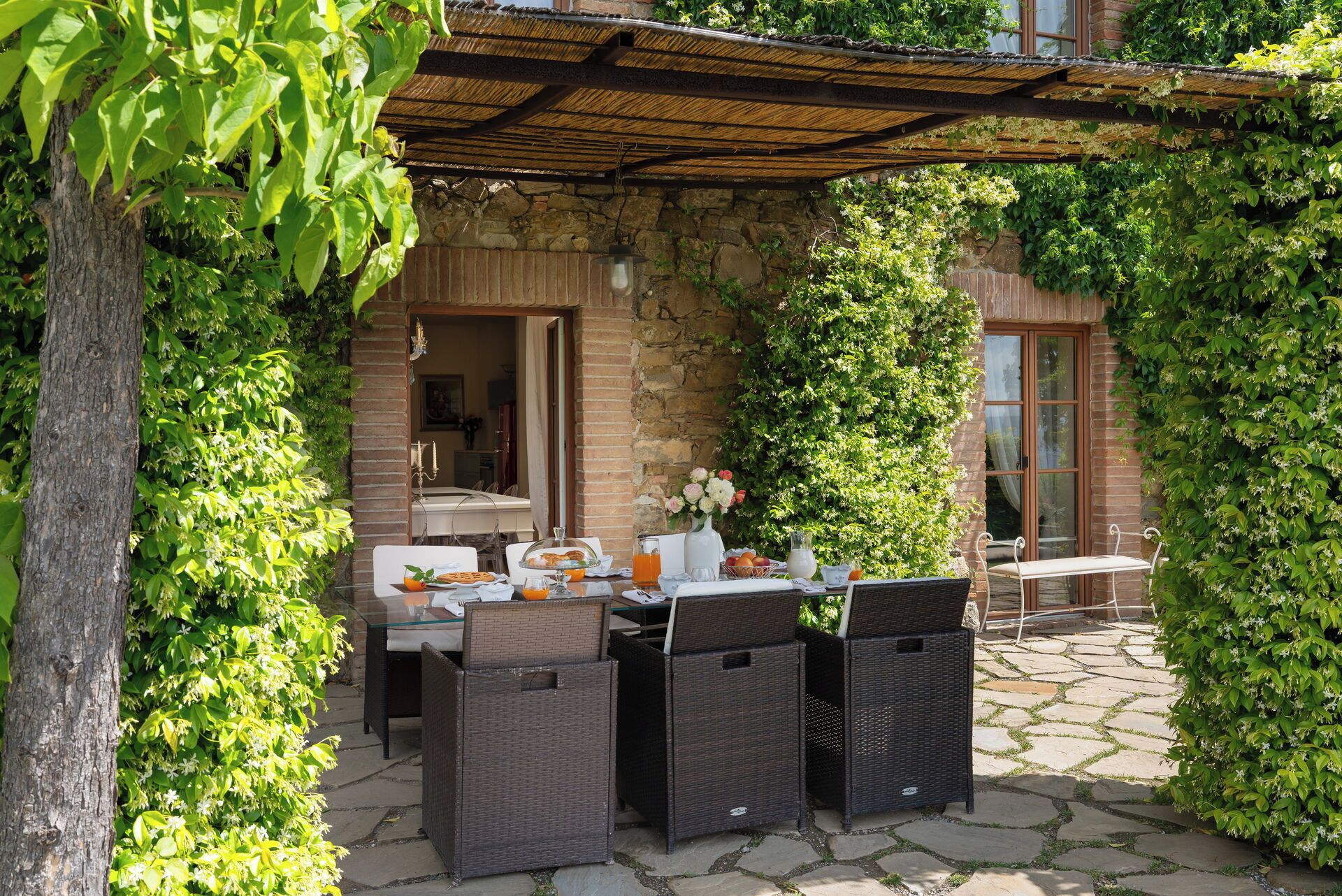 Un espace extérieur aménagé pour les repas, avec des meubles en osier, un mur en pierre et une vue sur l’intérieur.