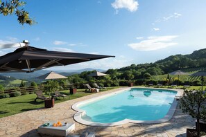 Piscina con sillas de descanso y sombrillas, rodeada de un patio de piedra y abundante vegetación.