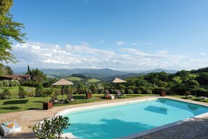 Ein Swimmingpool mit Liegestühlen und Sonnenschirmen, umgeben von einer Steinterrasse und üppiger Bepflanzung.