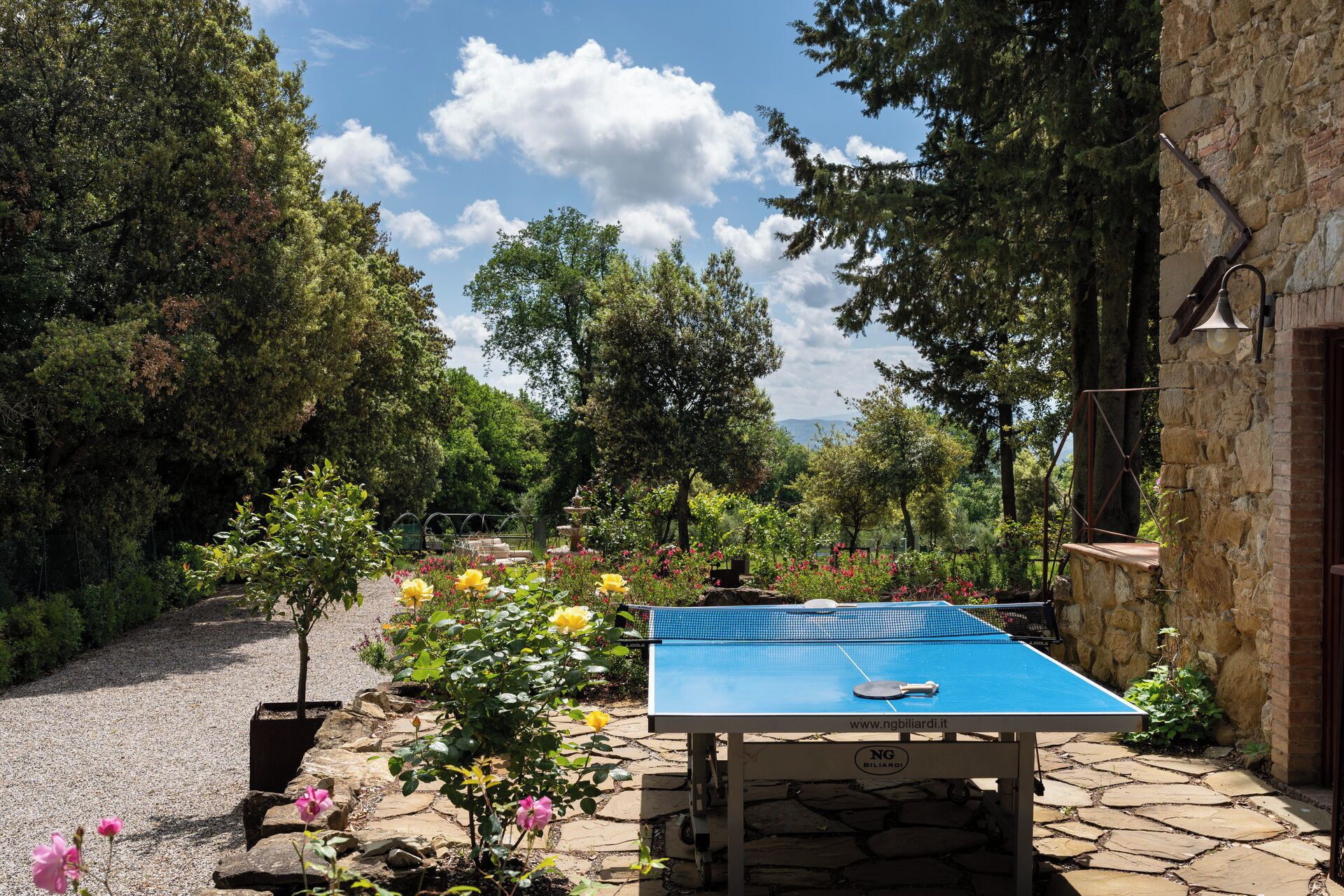 An outdoor ping pong table set up in a garden with stone pathways, surrounded by trees and flowers.