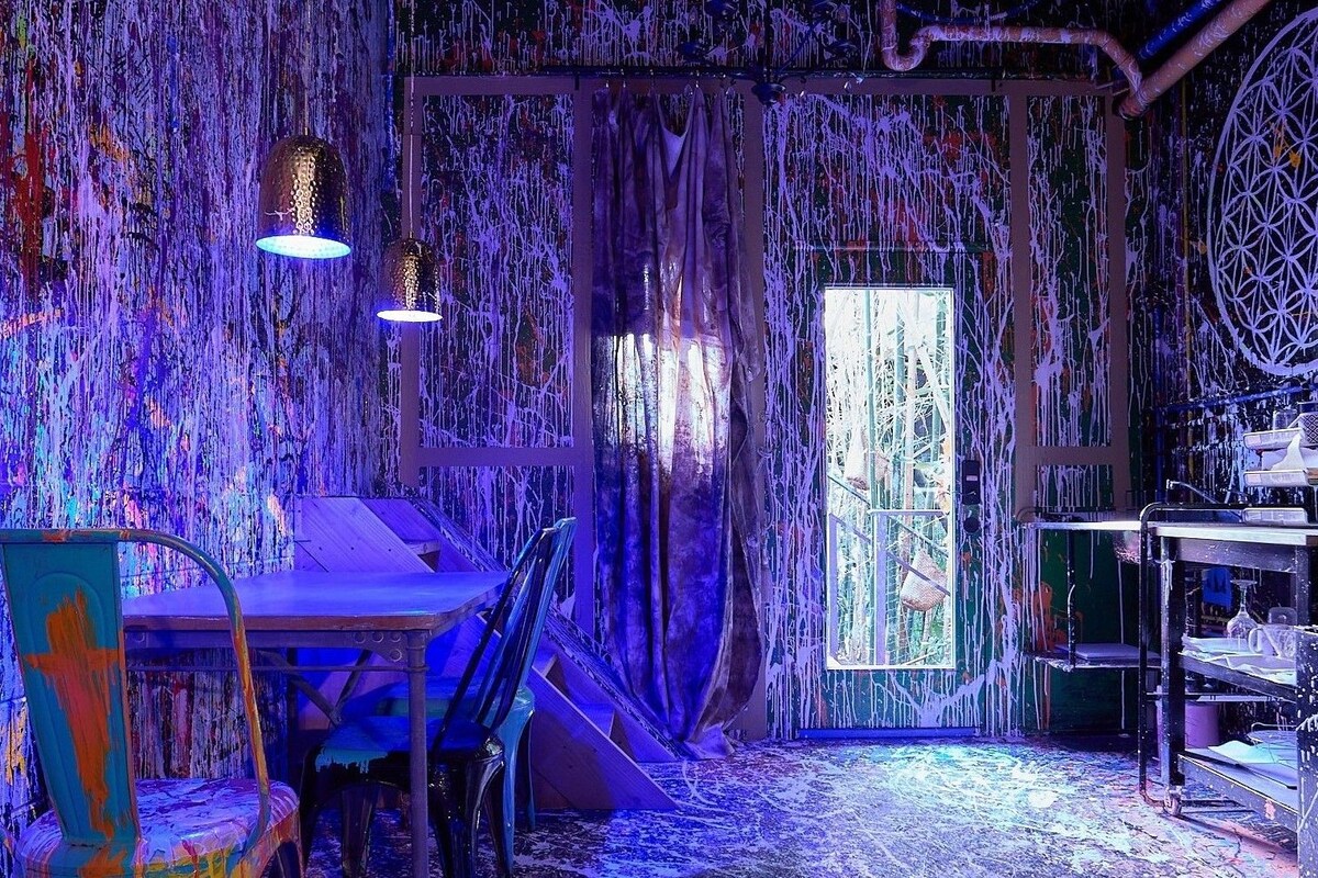 A room with a rustic aesthetic, featuring a wooden table, chairs, and a window with curtains.