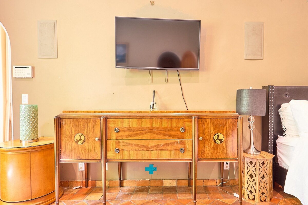 Habitación de hotel con tocador de madera, mesita de noche con lámpara, cama con ropa blanca y televisor de pantalla plana montado en la pared.