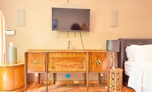 A hotel room with a wooden dresser, a bedside table with a lamp, a bed with white linens, and a flat-screen TV mounted on the wall. - Beverly Hills Spanish (Beverly Hills)
