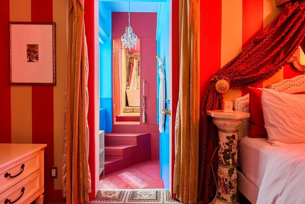 A room with a bed, a bedside table, a nightstand, a mirror, a framed picture on the wall, and a staircase leading to another room. - Beverly Hills Spanish (Beverly Hills)