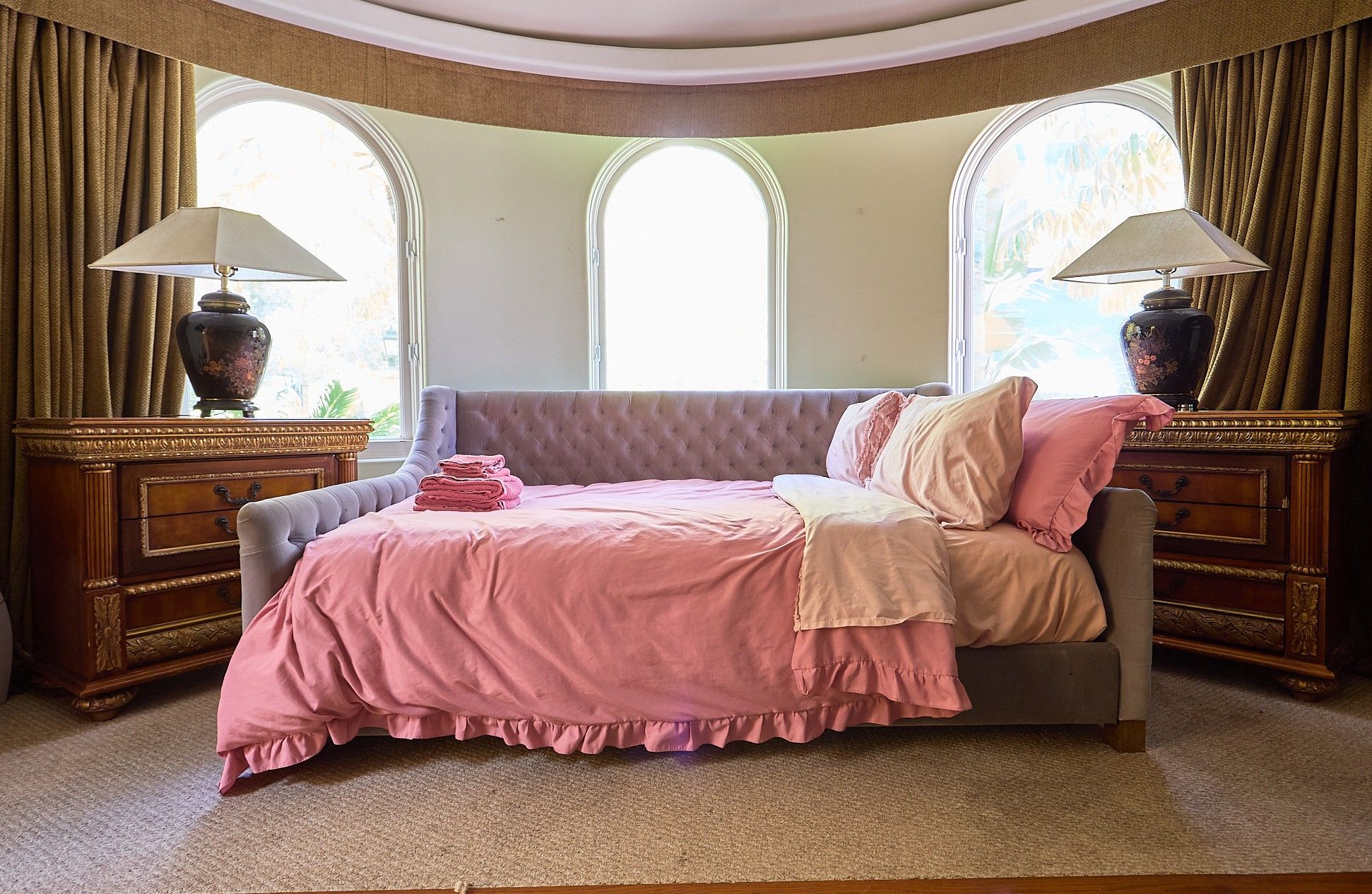A bedroom with a tufted headboard, a bed with pink bedding, two nightstands with lamps, and a large window with curtains.