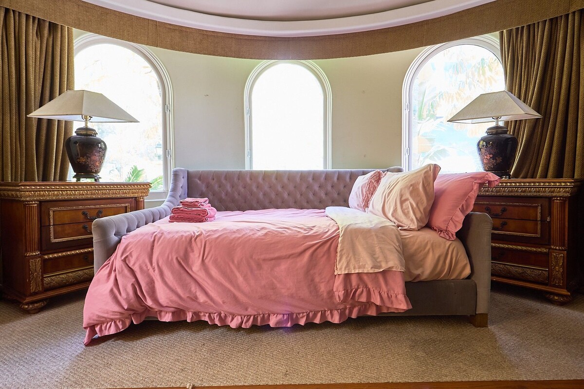A bedroom with a tufted headboard, a bed with pink bedding, two nightstands with lamps, and a large window with curtains.