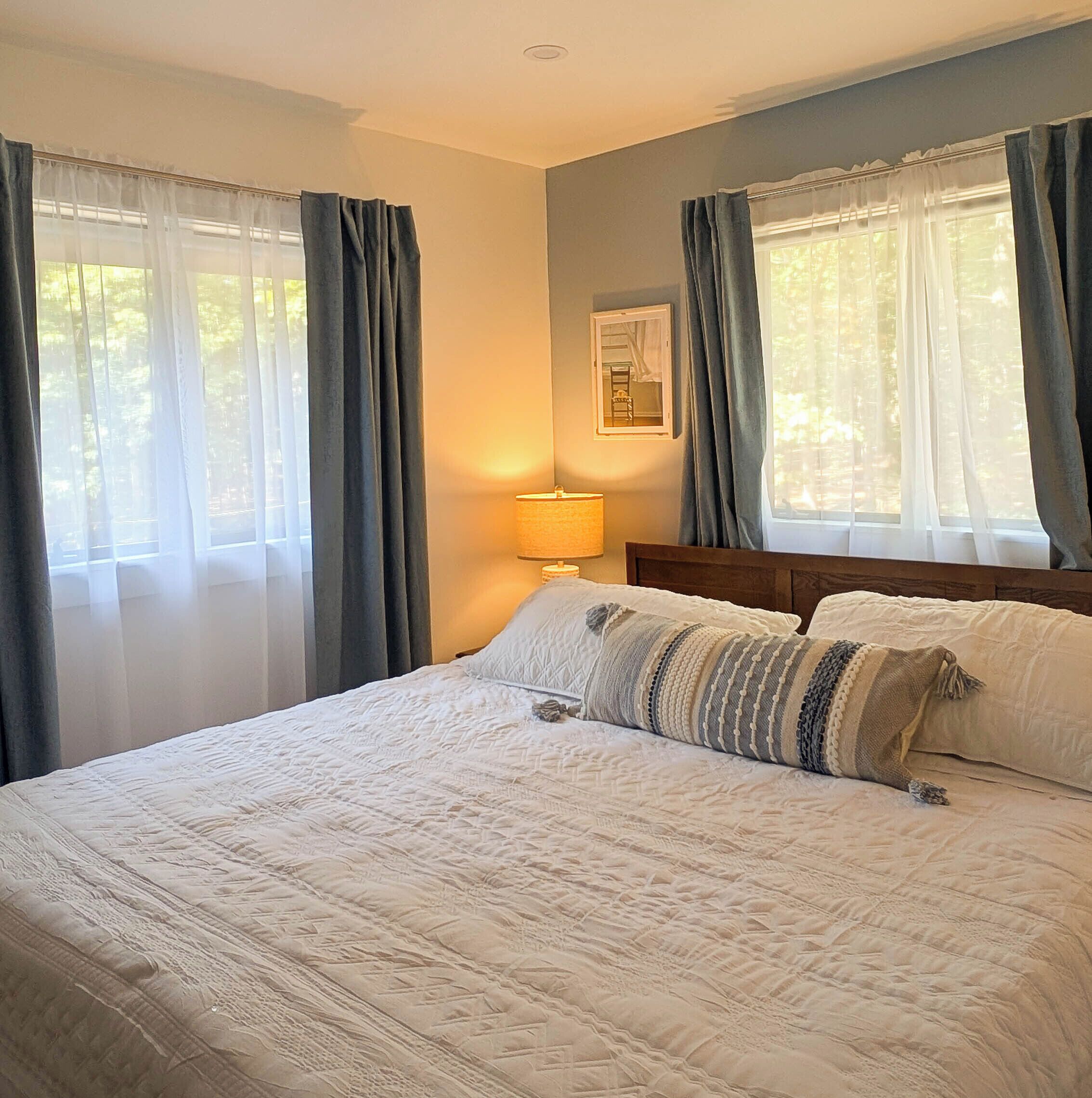 A bedroom with a bed, a nightstand, a lamp, and two windows with curtains.