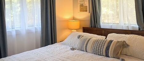 A bedroom with a bed, a nightstand, a lamp, and two windows with curtains.