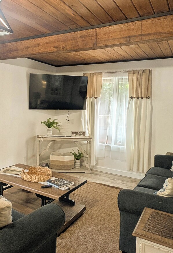 A living room with a wooden ceiling, a flat-screen TV mounted on the wall, a sofa, a coffee table, and a window with curtains. - Beautiful Bar Harbor House (Bar Harbor)