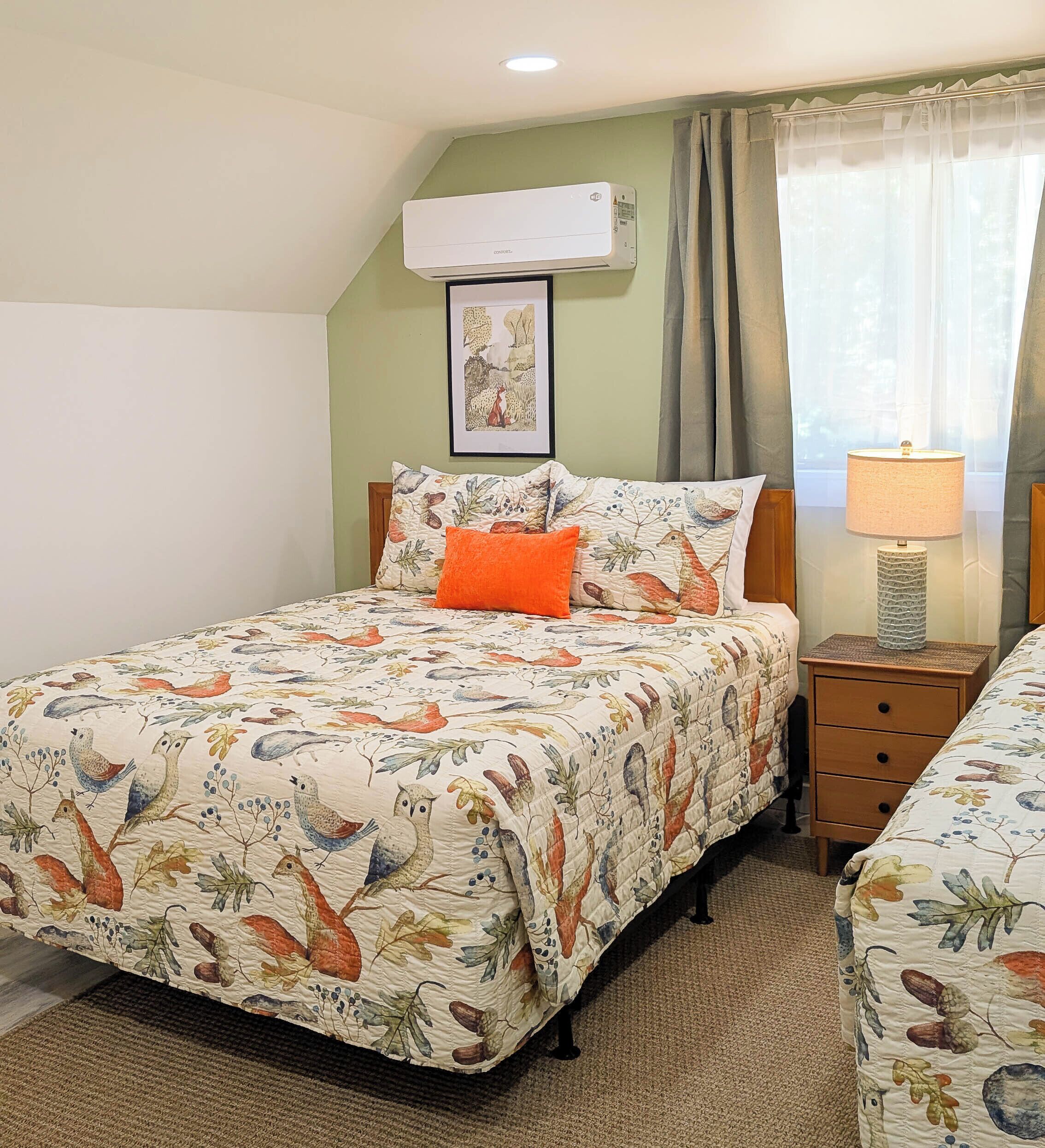 A bedroom with a bed featuring a floral and bird pattern, a bedside lamp, a framed picture on the wall, and an air conditioning unit.