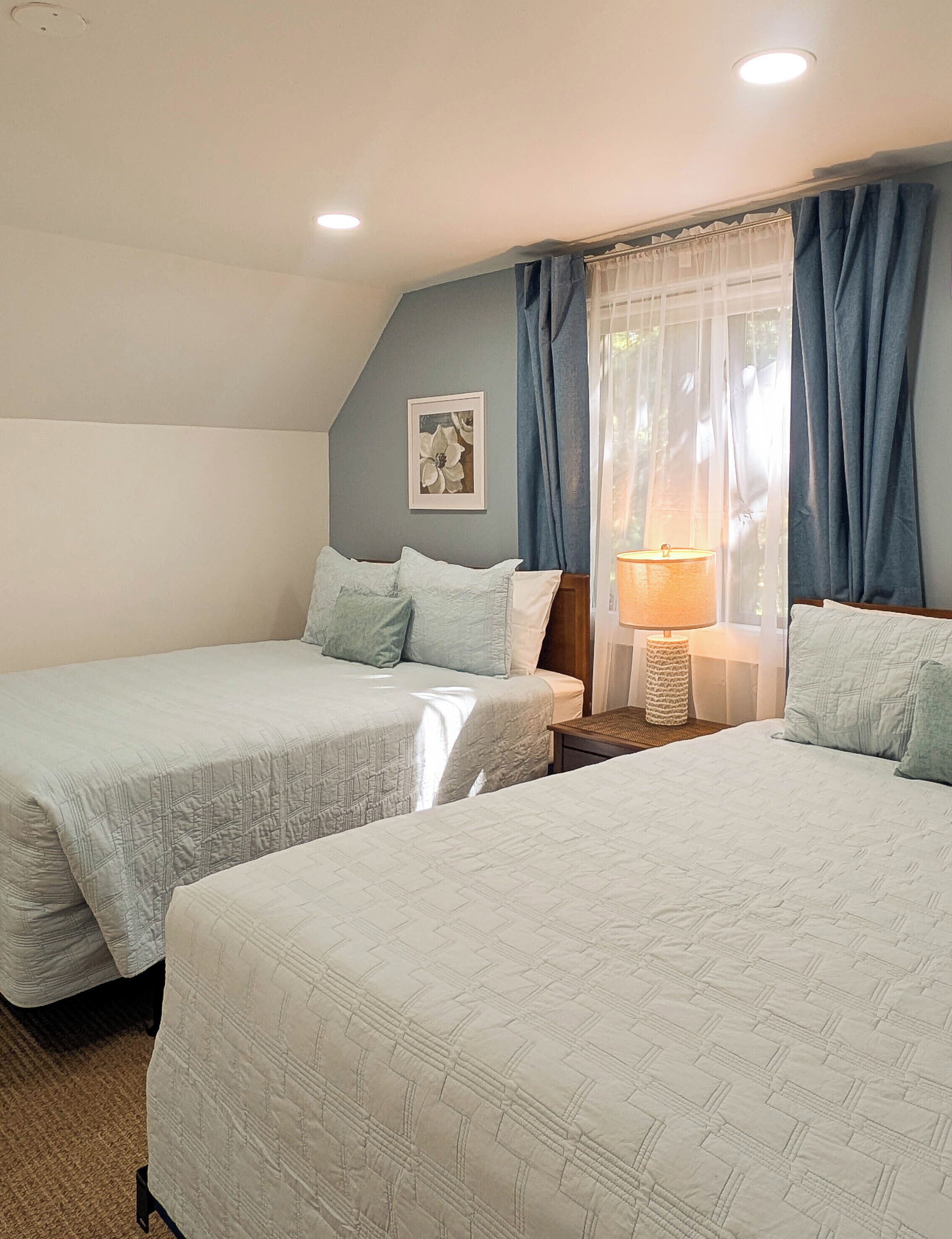 A bedroom with two beds, a nightstand with a lamp, and a window with curtains.