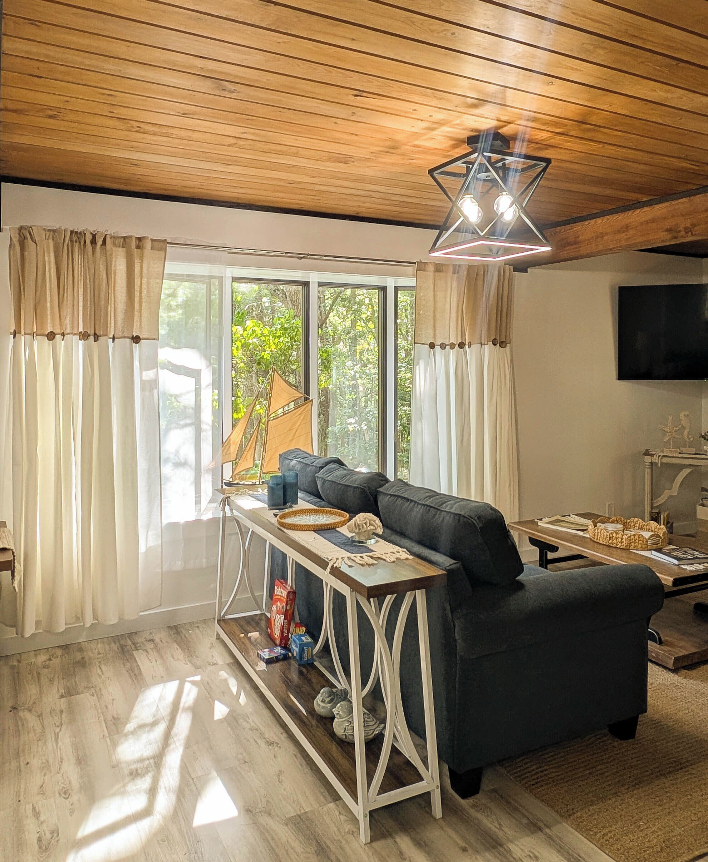 A modern living room with a black sofa, wooden ceiling, and a large window with curtains.