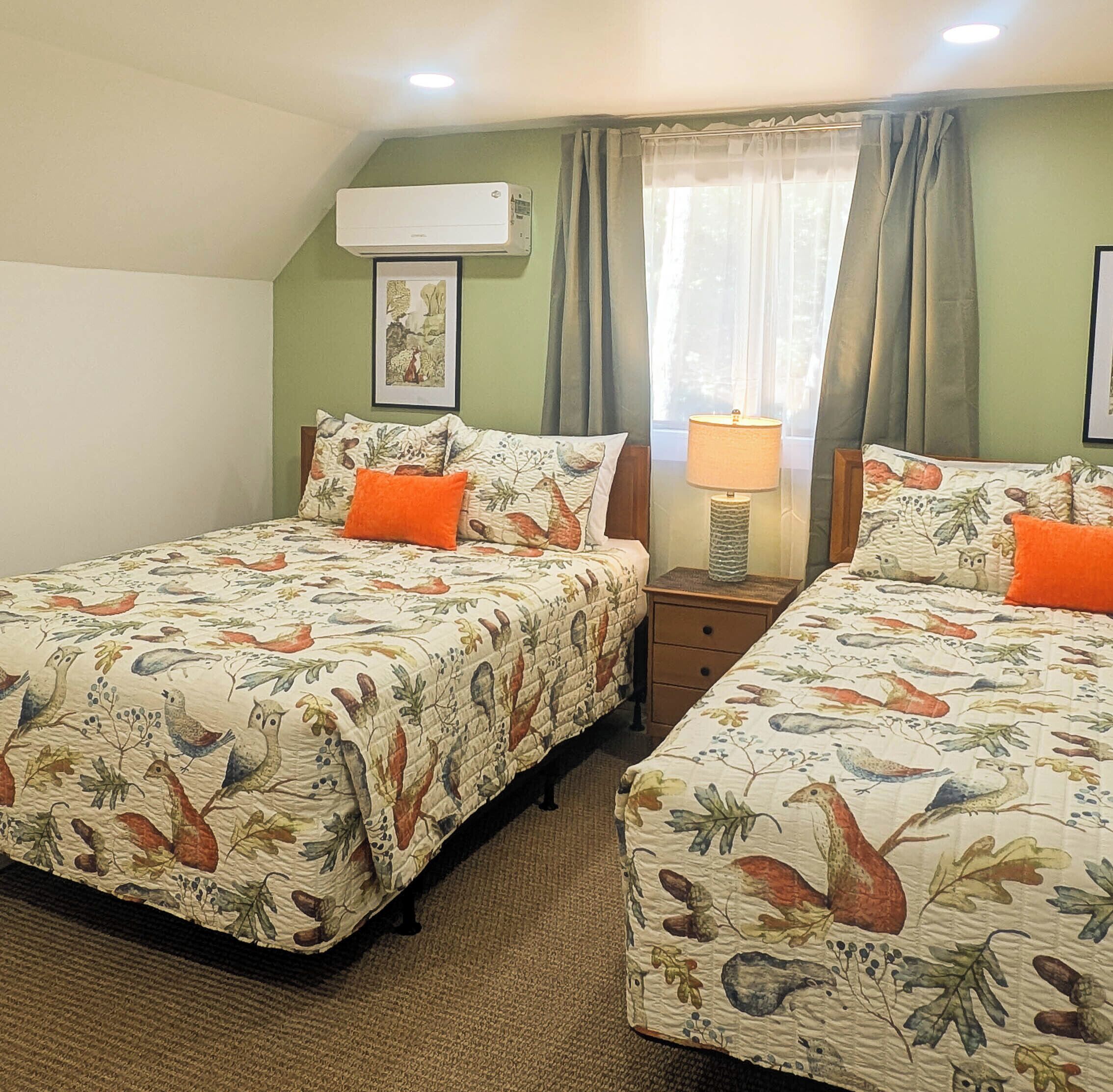 Un dormitorio con dos camas, cada una con ropa de cama floral y almohadas naranjas. Hay un ventilador de techo, una ventana con cortinas y un cuadro en la pared.
