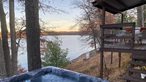 A hot tub with a view of a lake and trees, a wooden deck, and a covered seating area. - Rustic Lake Nokomis Cabin with Hot Tub, Trails, and Family Fun for 10! (Tomahawk)