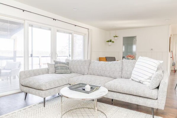 A modern living room with a grey sectional sofa, a small round coffee table, and a large window providing natural light. - Rustic Lake Nokomis Cabin with Hot Tub, Trails, and Family Fun for 10! (Tomahawk)