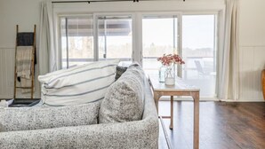 A living room with a sofa, a small table with a vase, and a view through large glass doors. - Rustic Lake Nokomis Cabin with Hot Tub, Trails, and Family Fun for 10! (Tomahawk)