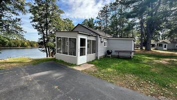 Une petite cabane avec une véranda grillagée, un barbecue et un banc situés près d’un lac.