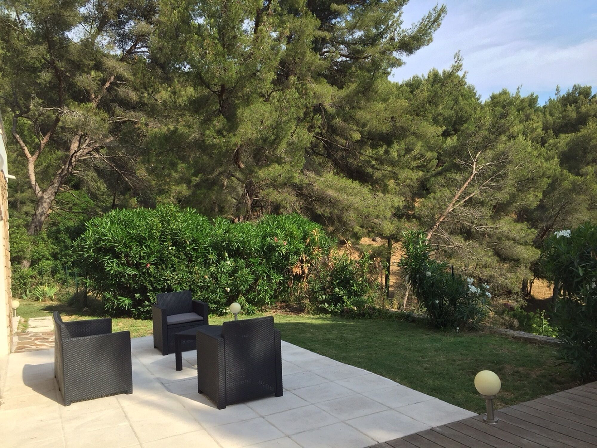 An outdoor patio area with a seating arrangement of three black wicker chairs and a table, surrounded by a well-maintained lawn and dense trees.