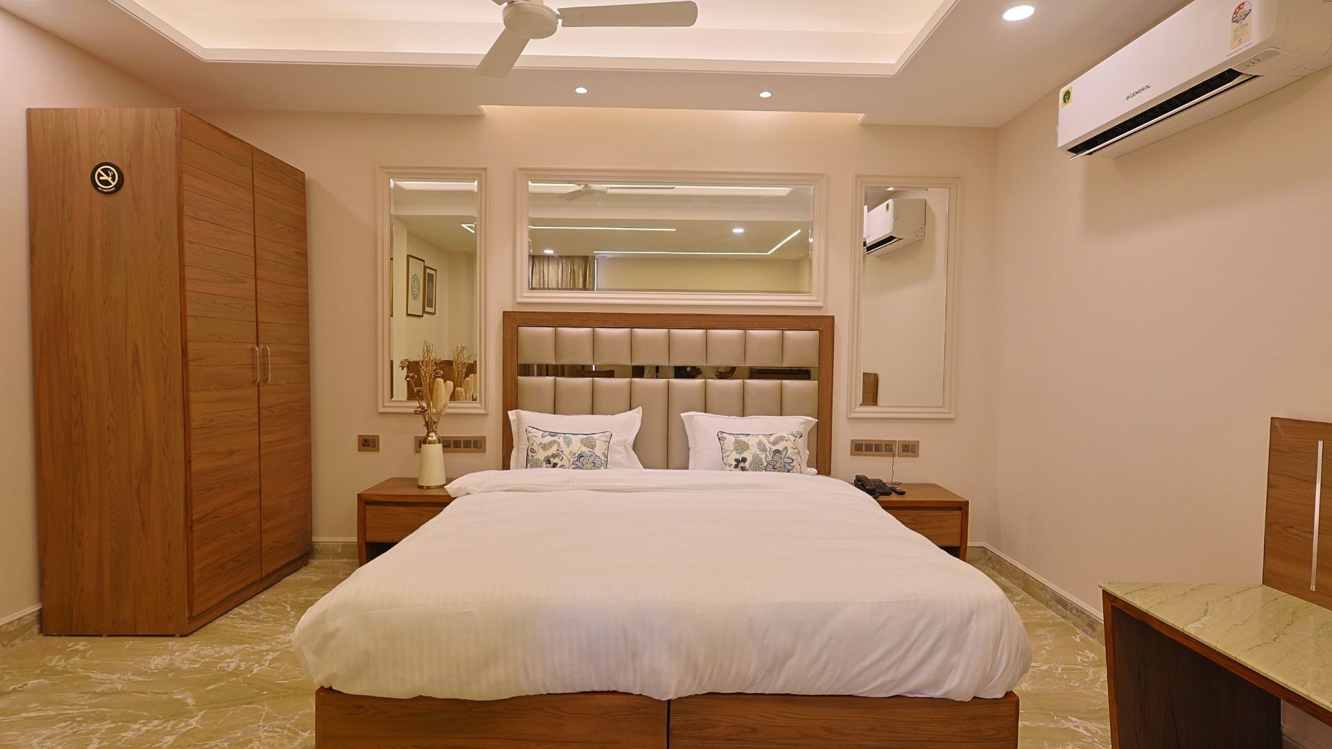 Un dormitorio con una cama grande, un armario de madera, dos mesitas de noche, un ventilador de techo y un equipo de aire acondicionado.