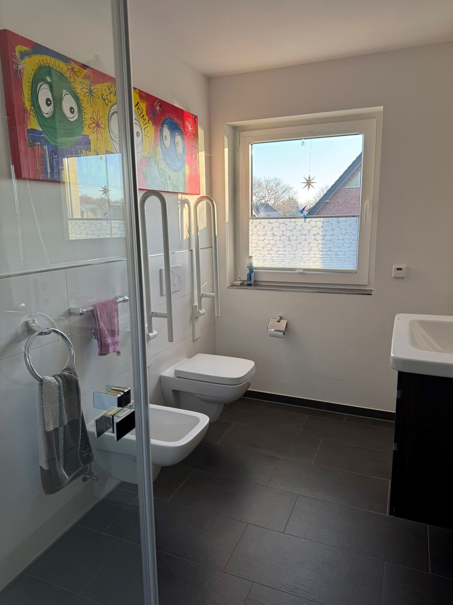 Ett modernt badrum med en duschkabin i glas, en toalett, en bidé och ett fönster med utsikt över ett hus.