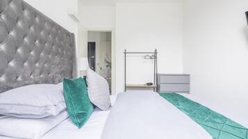 Une chambre à coucher avec une tête de lit capitonnée, un lit recouvert de draps blancs et d’un plaid vert, une table de chevet avec une lampe, et un présentoir en métal adossé au mur.